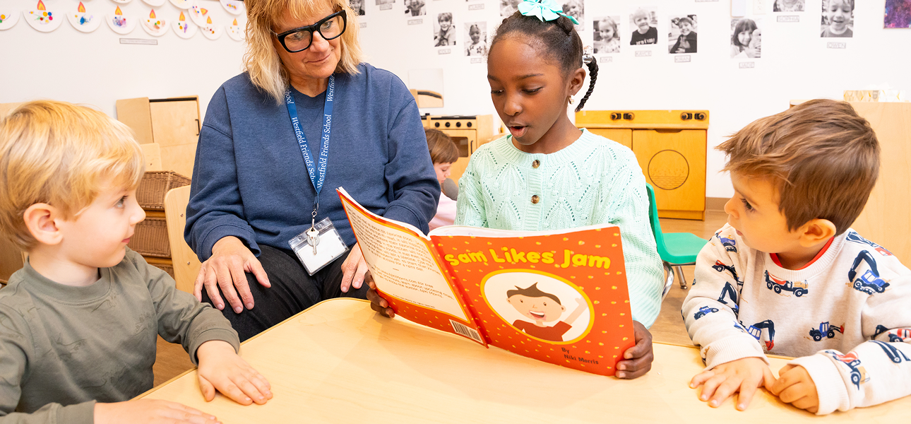 A young girl reads a book titled 'Sam Likes Jam' aloud to two younger children and an adult teacher sitting around a low classroom table.