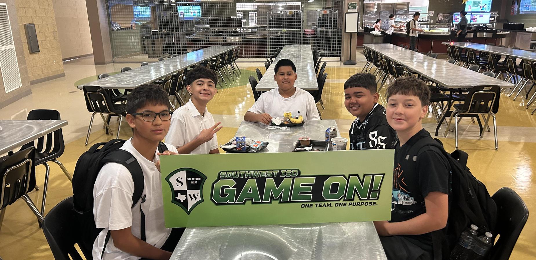 Five students pose at a cafeteria table holding a sign that reads 'GAME ON!'