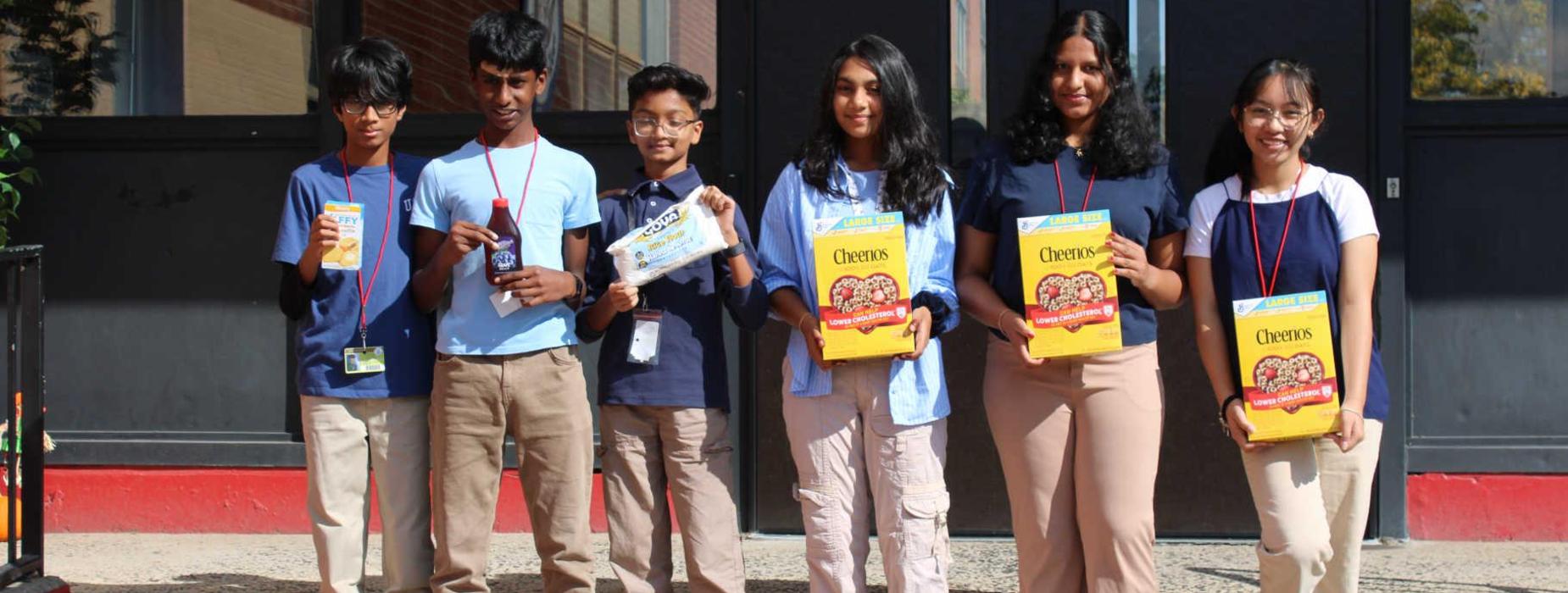 Six students holding cereal boxes and snacks outside a building.