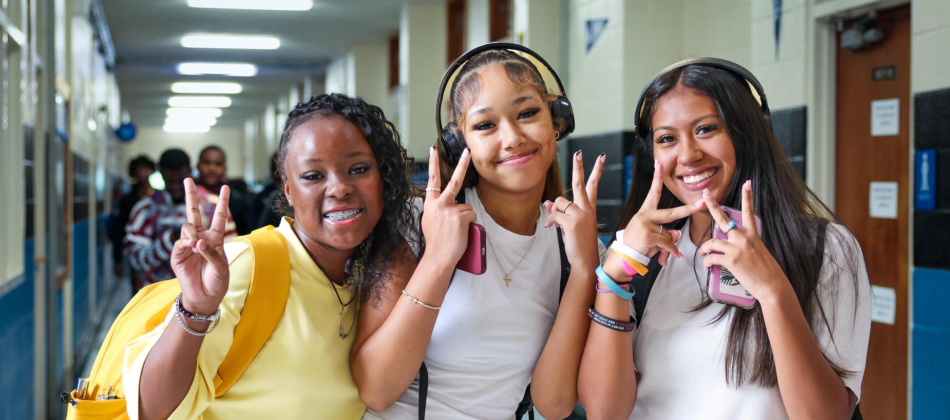 Three young women pose together, smiling and making peace signs in a school hallway.