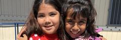 Two smiling girls embracing, standing outdoors in bright sunlight.