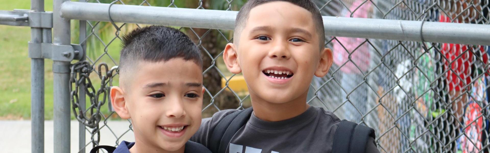 Two boys standing together, smiling, outside near a fence.