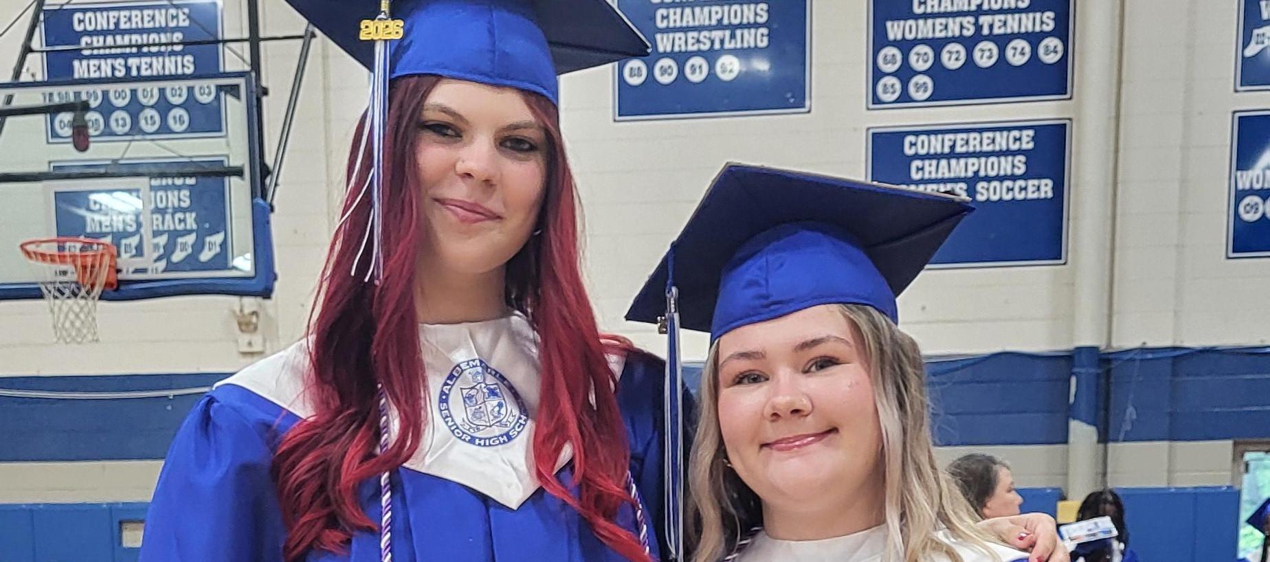 Two graduates in blue gowns stand side by side, showcasing their attire and tassels.