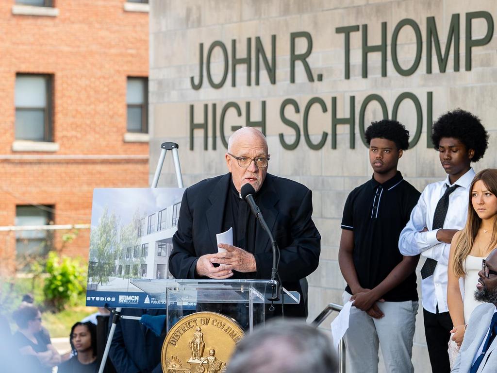 A man speaks at a podium outside John R. Thompson High School, with students and a rendering behind.