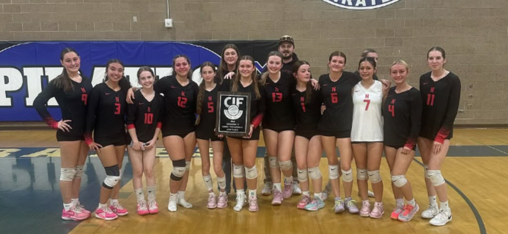 Team of volleyball players in black uniforms holding a championship plaque while posing on the court.