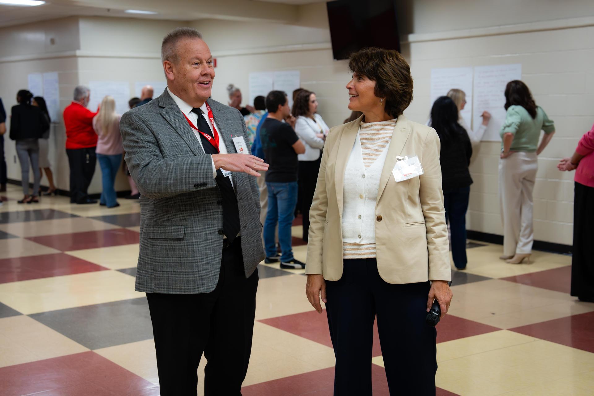 Two people in conversation in a hall with others in the background.