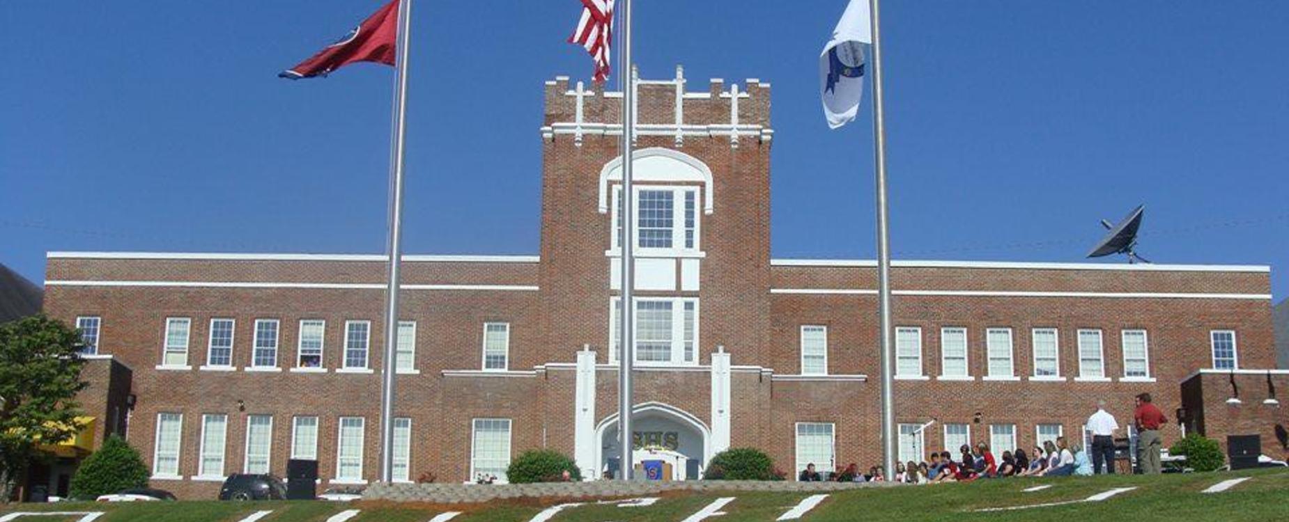 A brick building with flags in front and people sitting on the lawn.