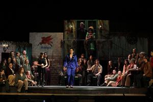 girl in blue suit singing in middle of stage while cast in rugged clothes watches