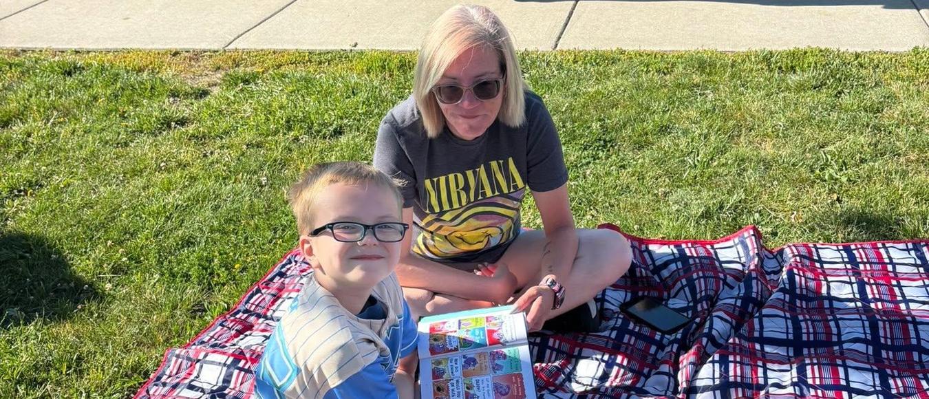 A woman and a young boy sit on a plaid blanket on the grass together reading a book.