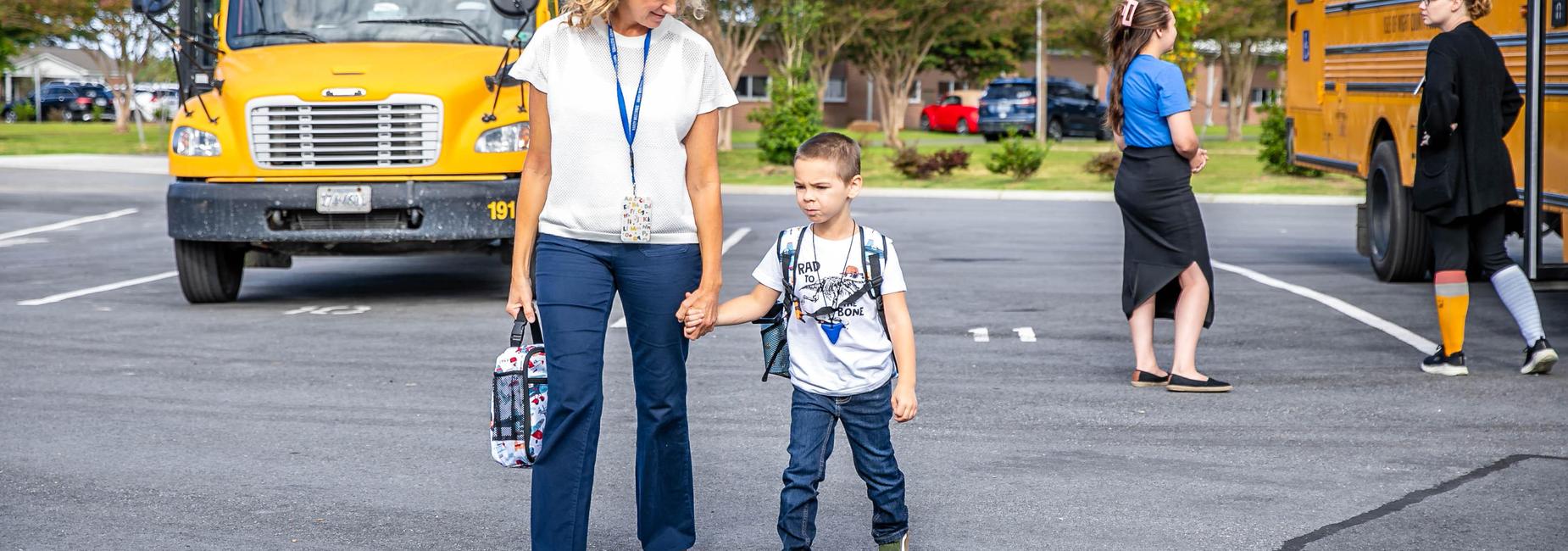 Teacher holding a child's hand while walking past a school bus in a parking lot.