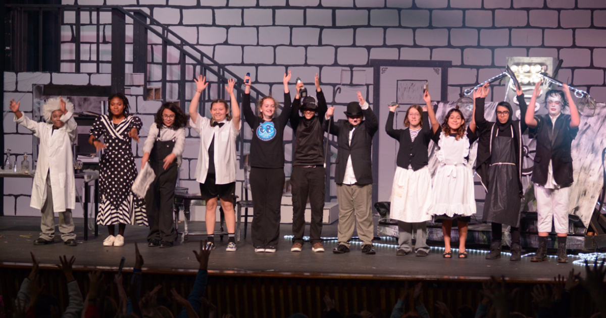 Middle and high school students take a bow on stage during their production of The Misfortunes of Charlie Chaplin. The cast, dressed in costumes ranging from lab coats to period attire, stands in a line with their arms raised. The backdrop features a black-and-white brick design, evoking a vintage setting. Enthusiastic audience members can be seen reaching up from the foreground.