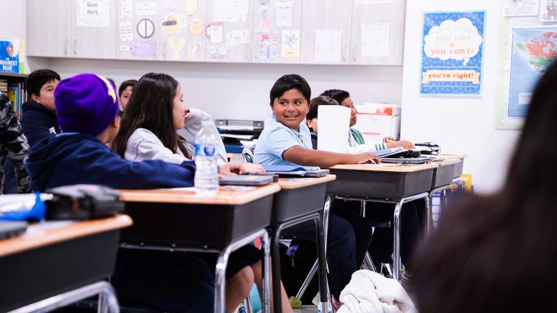 Group of students in a classroom, one boy smiling while looking back at others.