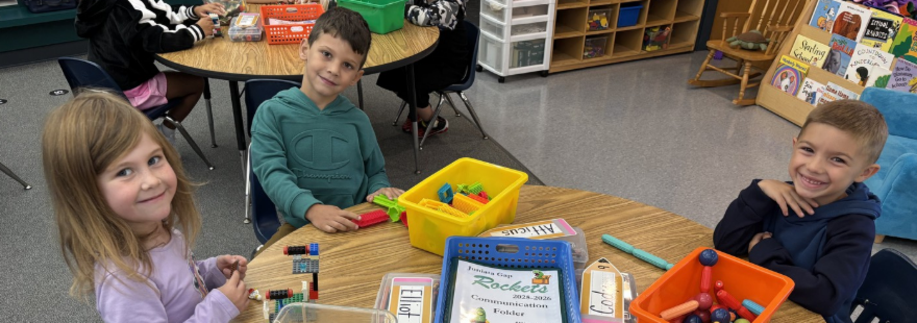 Three kids smiling at a table with building blocks and organizational boxes.