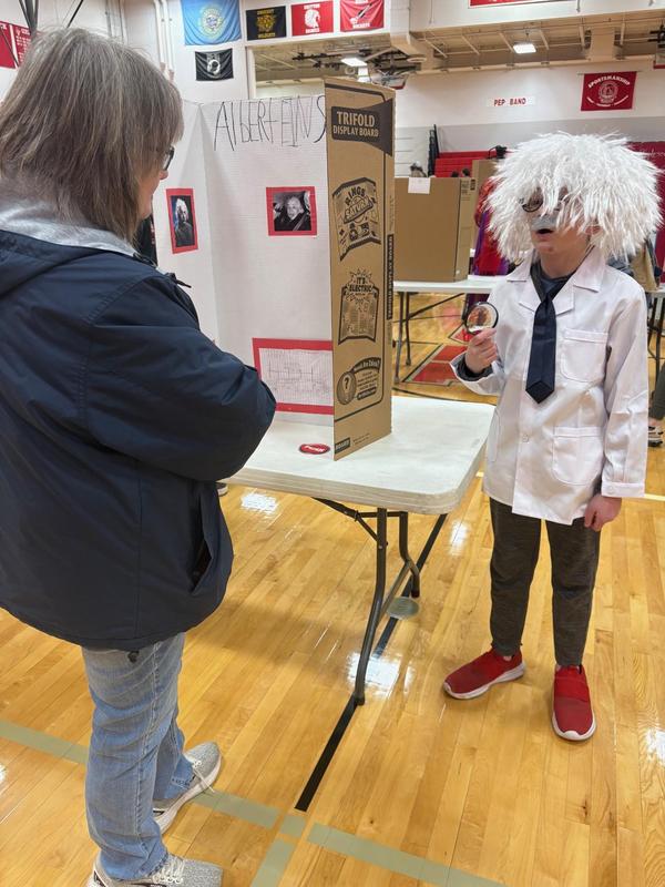 A child dressed as Einstein presents a project to an adult at a school event.