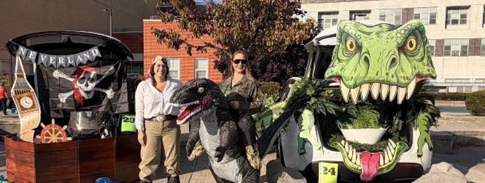 Two women in costumes pose with dinosaur-themed cars at a festive outdoor event.