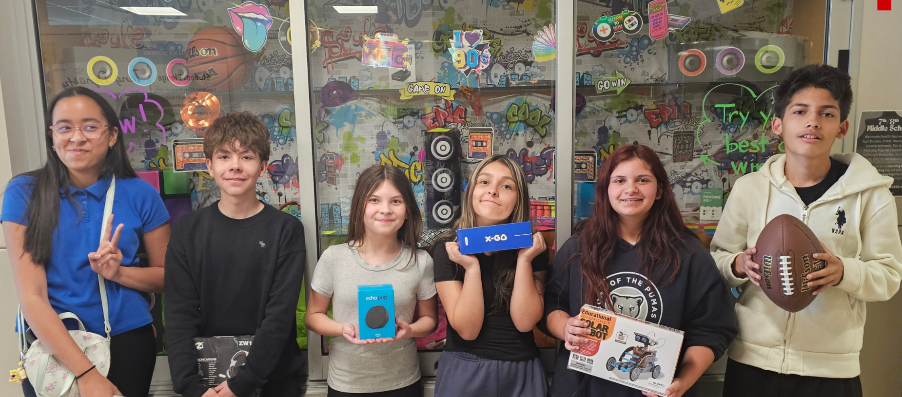 A group of six young people posing with prizes in front of a colorful display.