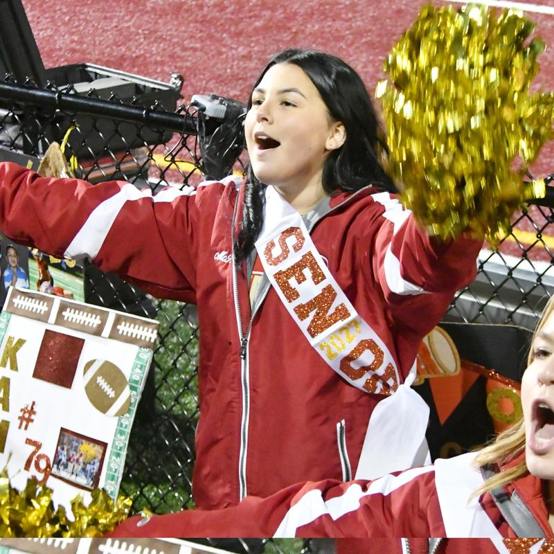 Cheerleader in a red jacket celebrates with pom-poms at a football game.