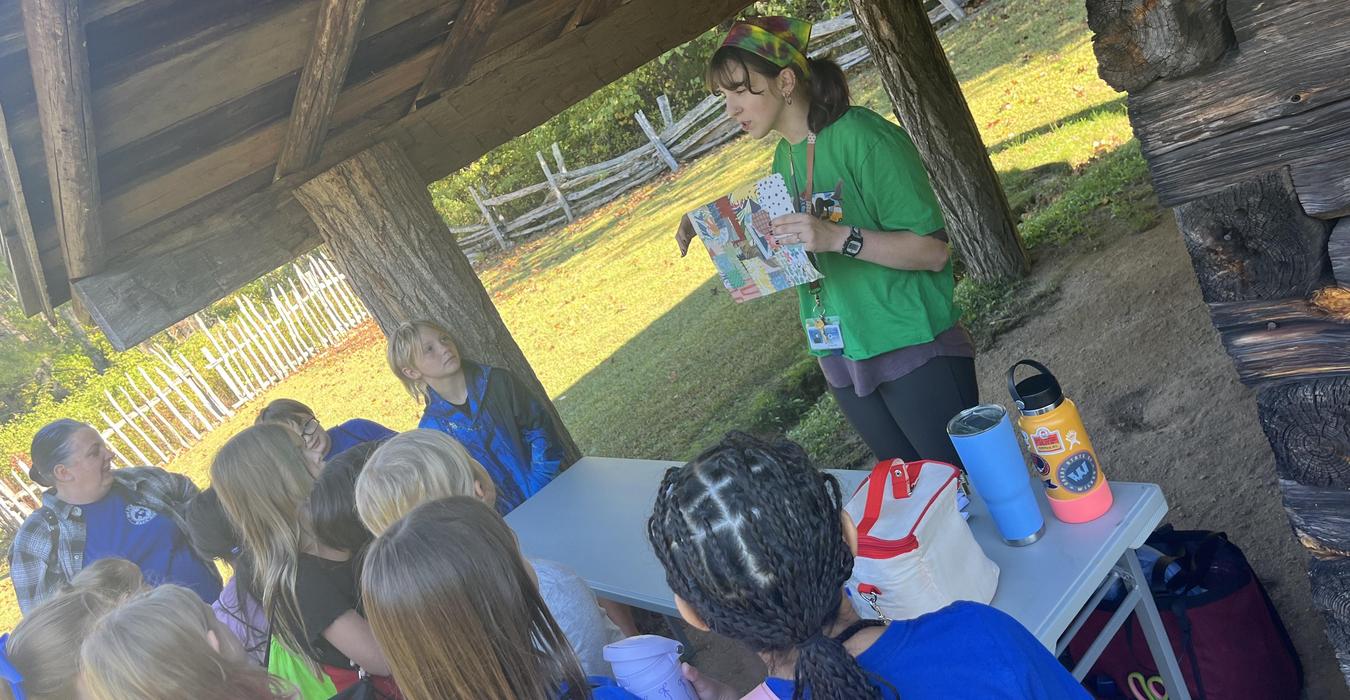 Teacher presenting to a group of children outdoors under a shelter.