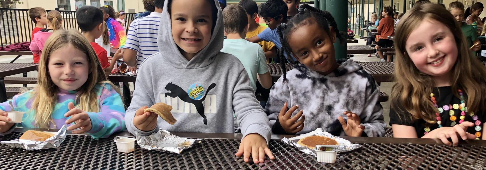 Four elementary school students eating pancakes at an outdoor picnic table