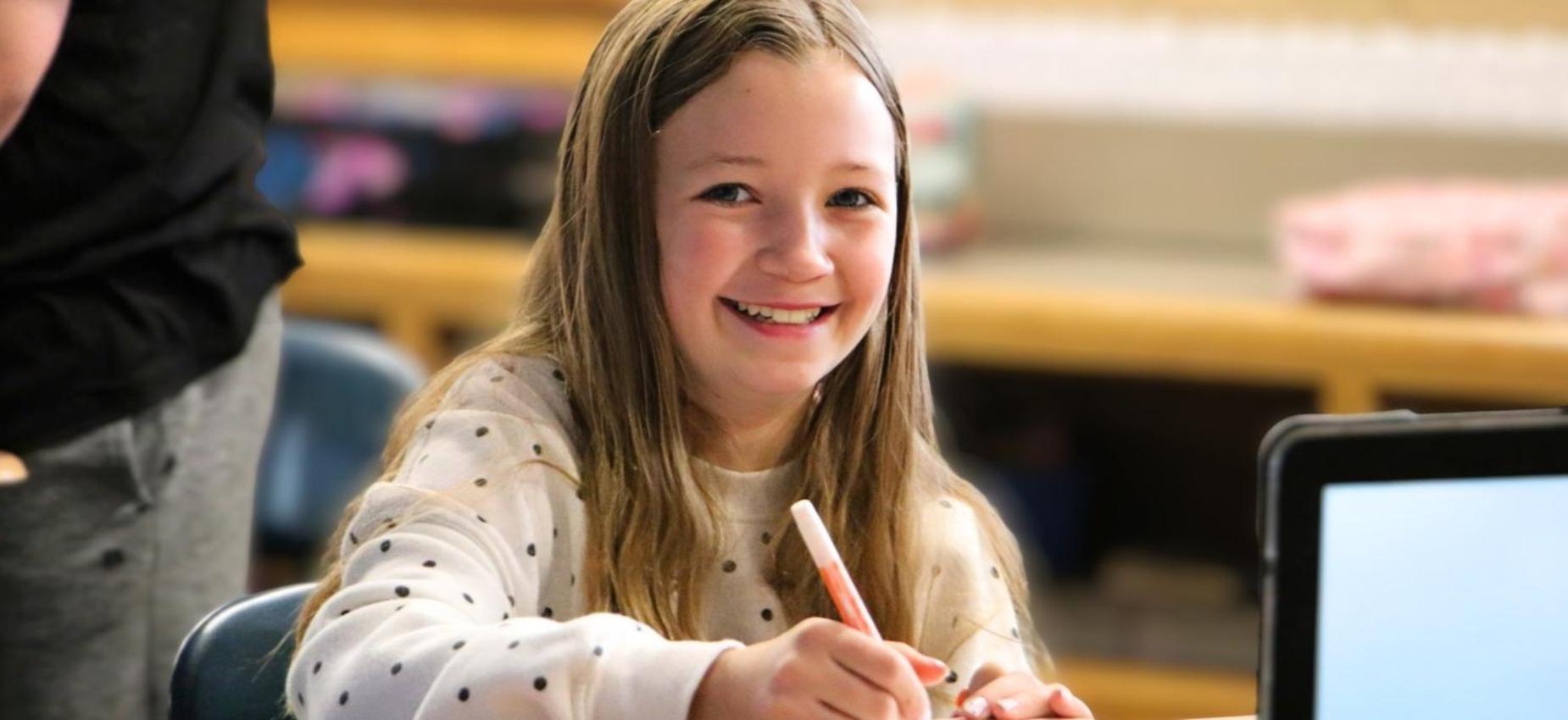 Smiling girl holding a marker while sitting at a desk.