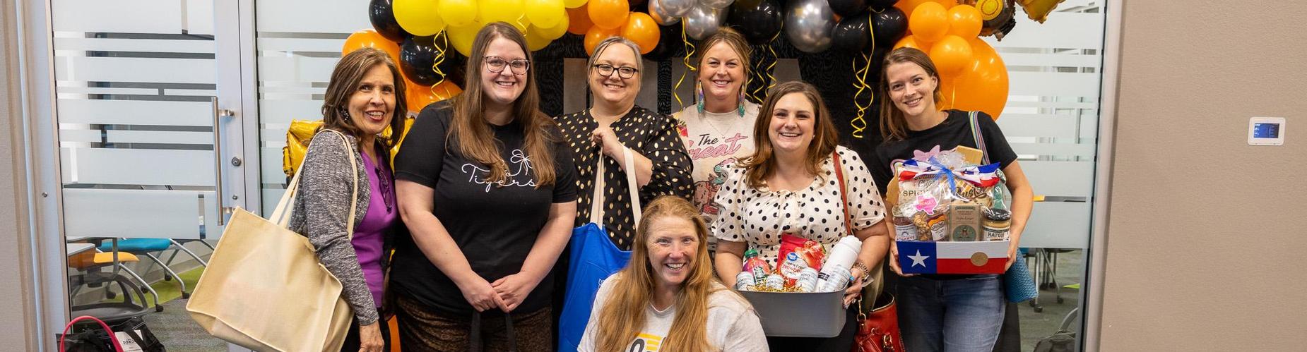 Group of seven people smiling together in an office with balloons and gifts.
