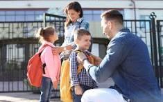 A parent assists two children with their backpacks outside a school.