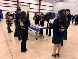 FFA students wearing navy blue jackets with gold lettering gather around a table set up for judging inside a large indoor facility. Each student is holding a clipboard, focused on evaluating items displayed on the table as part of an agricultural competition or practice session.