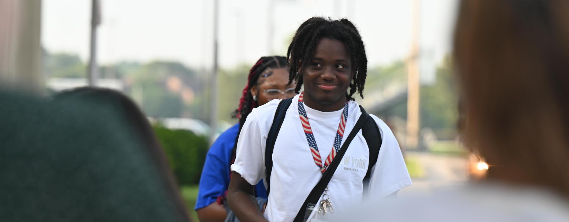 A young man with dreadlocks smiles while wearing a lanyard, with others in the background.