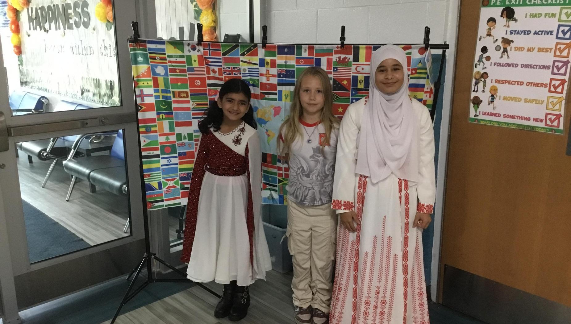 Three girls standing in front of a colorful flags backdrop in a school hallway.