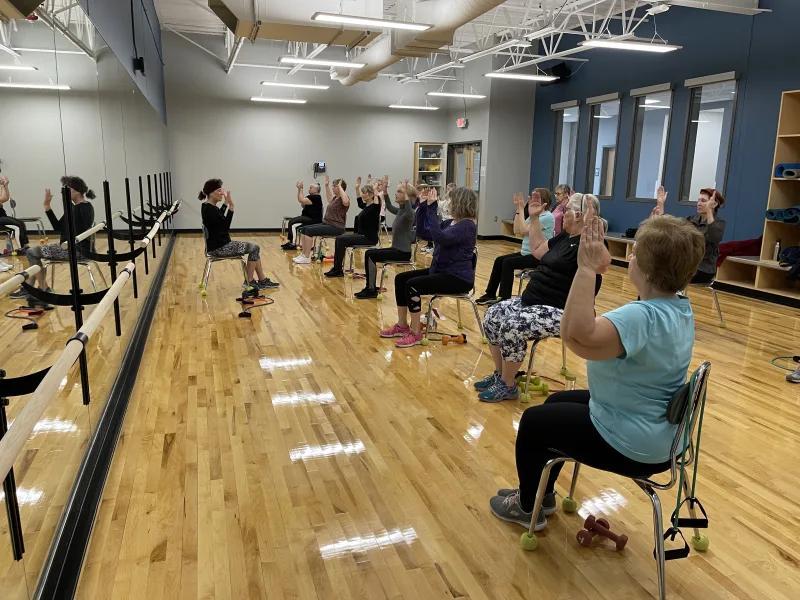 Group fitness class with participants in a dance studio sitting in chairs facing an instructor