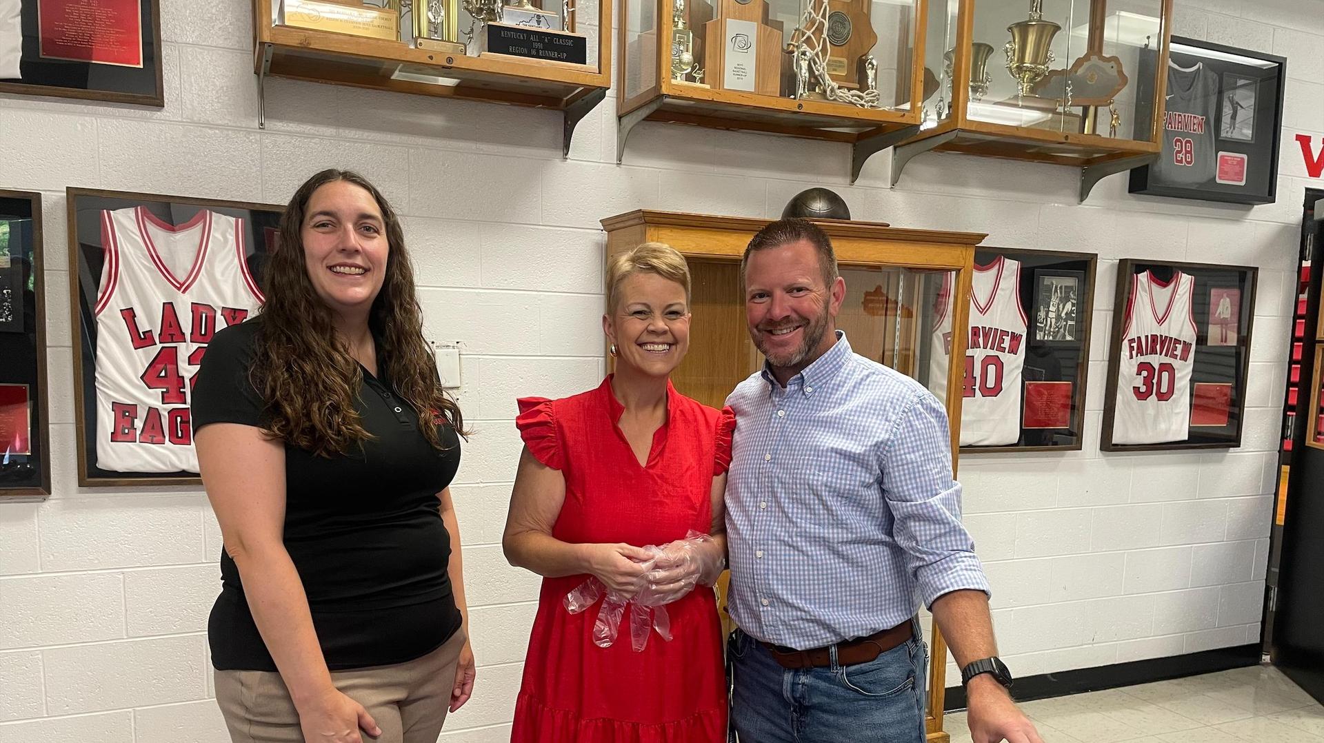 Three people posing together in a school gym, smiling and dressed nicely.
