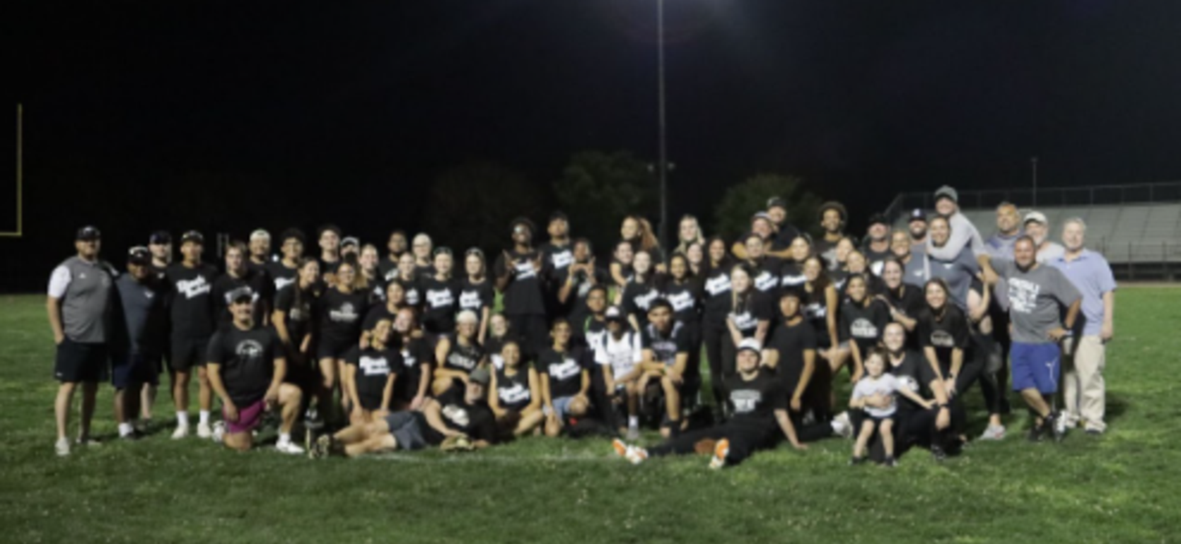 Group photo of a large team on a football field at night, wearing matching shirts.