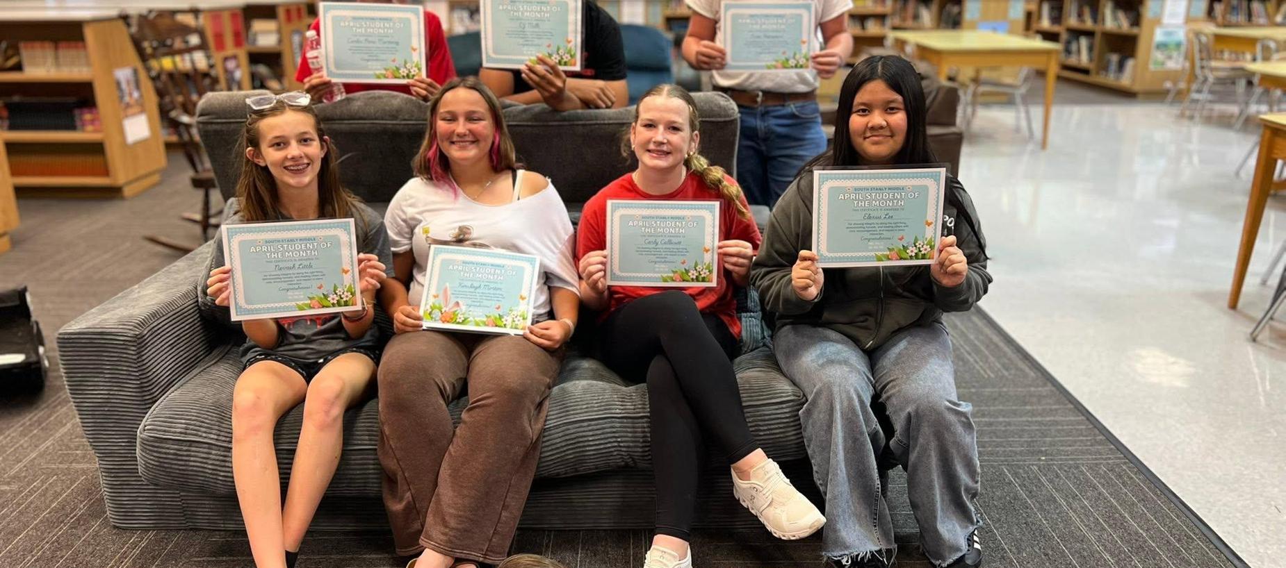 Group of girls holding certificates, seated on a couch in a library.