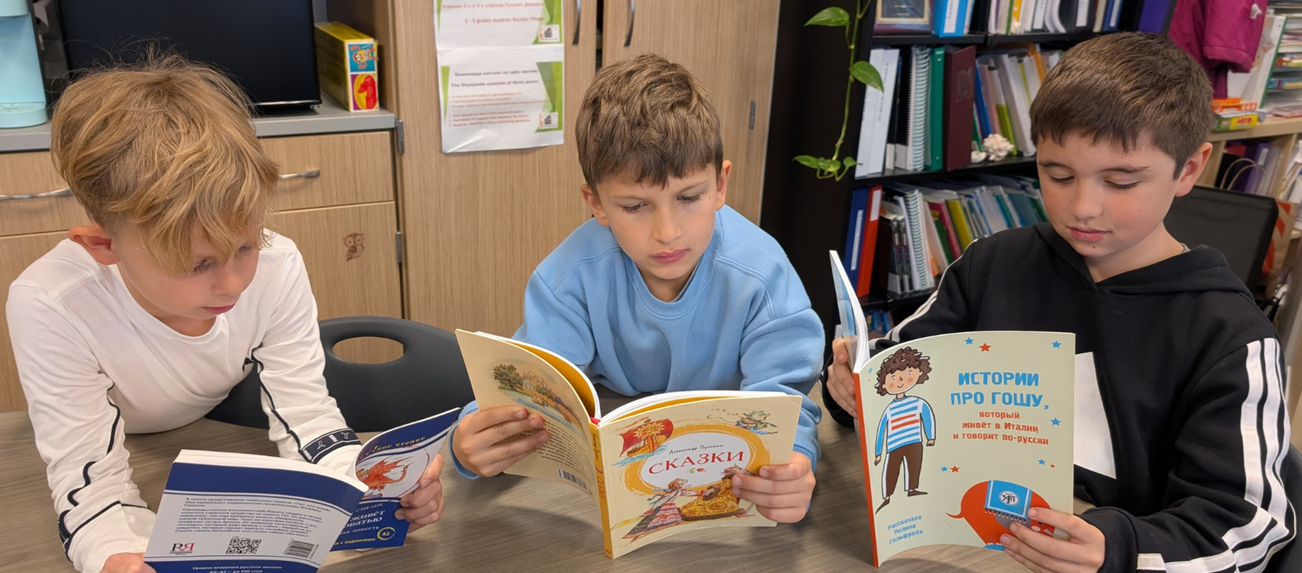 Three children reading books at a table, focused on their individual texts.