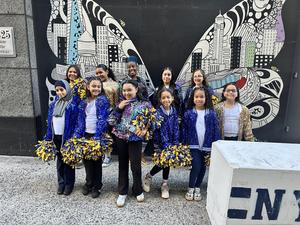 A group of cheerleaders in sparkly jackets poses in front of a colorful mural.