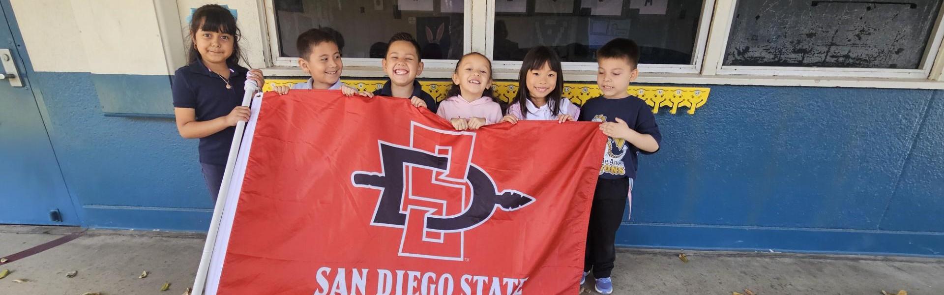Children joyfully holding a San Diego State University flag outdoors.