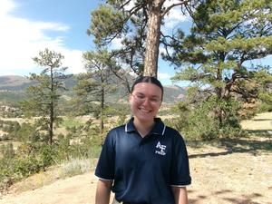 Meghan smiling in a navy blue Air Force Prep polo shirt in front of a scenic mountain landscape.
