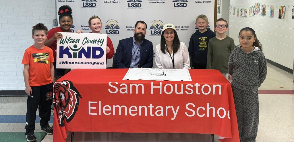 kids standing around a table that says Sam Houston Elementary School and holding up a sign that says Wilson County Kind with two adults sitting at the table in front of a wall that has the LSSD logo on it