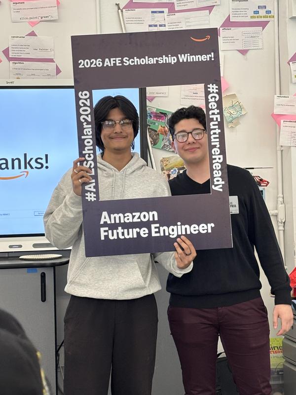 Two students pose with a scholarship frame in a classroom setting.