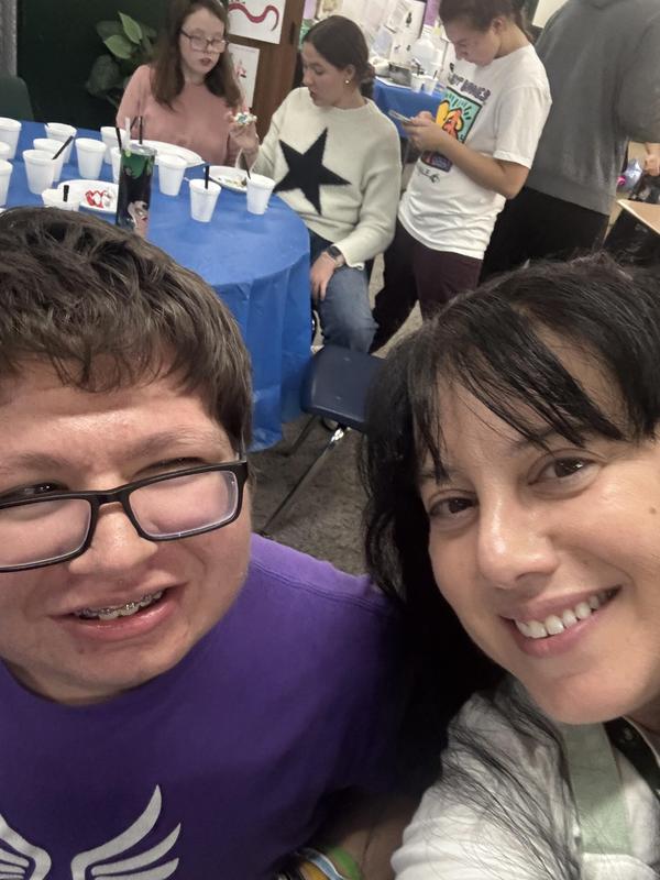 A close-up of two people smiling, with a classroom background and snacks on the table.