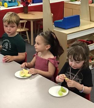Three children seated at a table enjoying slices of apple on white plates.