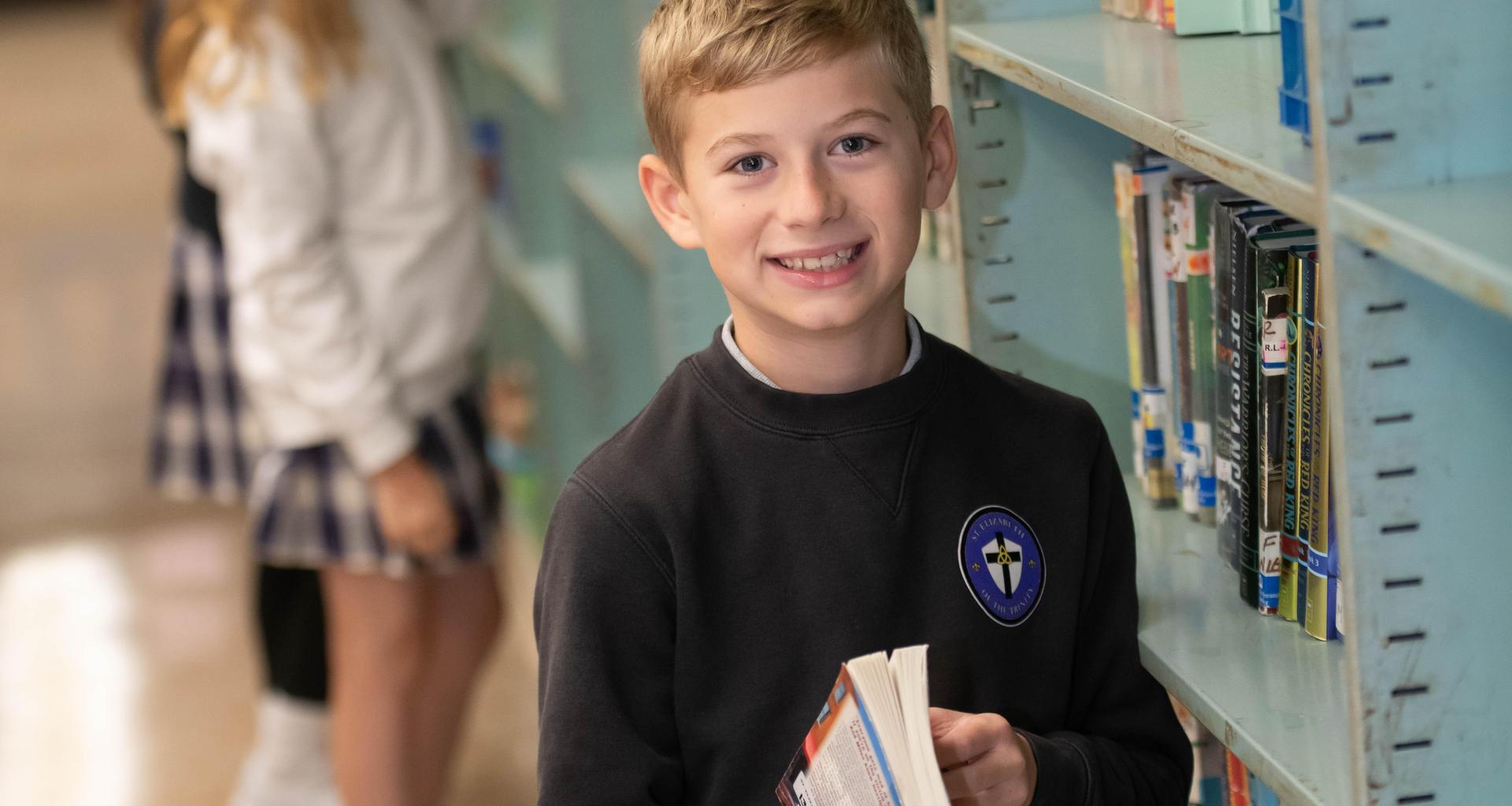 Smiling boy holding a book in a library, surrounded by shelves.