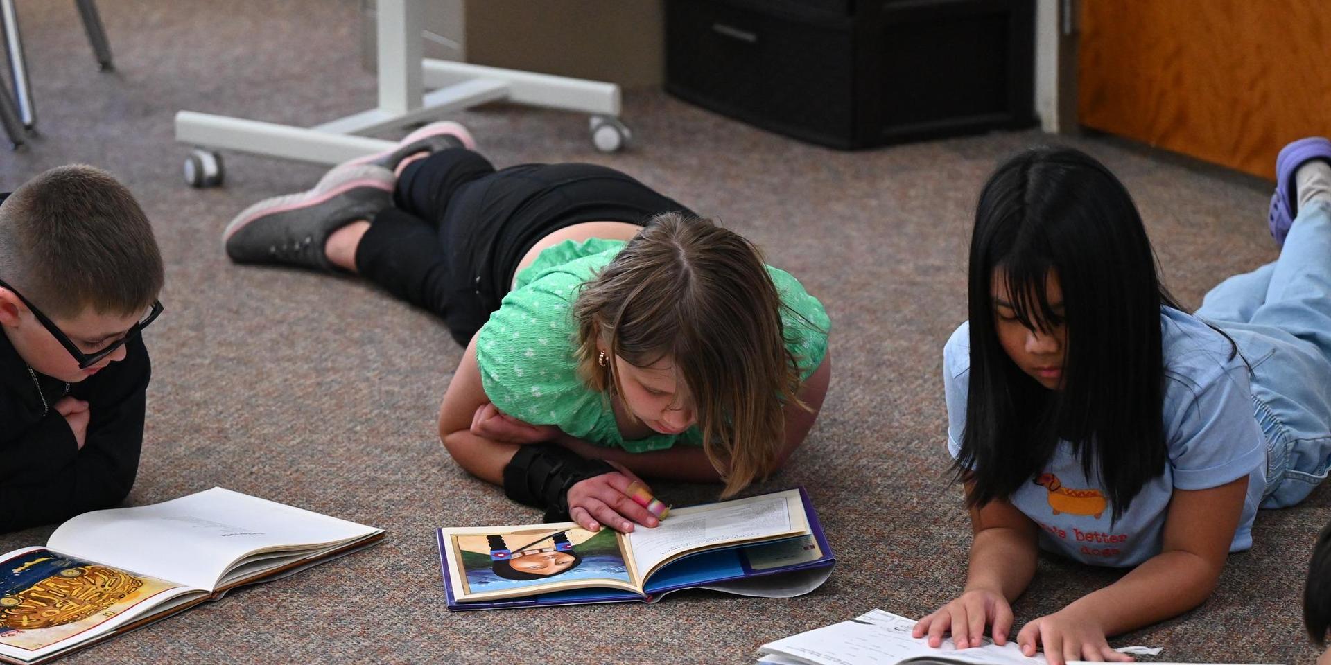 Three children lie on the floor reading books, focused and engaged in their activity.