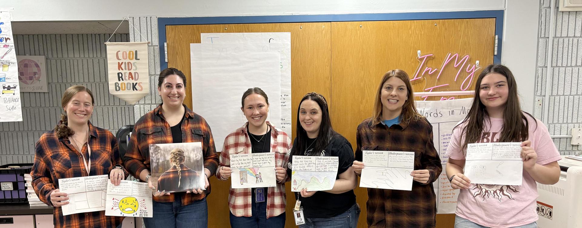 Six women holding posters in a classroom, wearing plaid shirts and smiling.