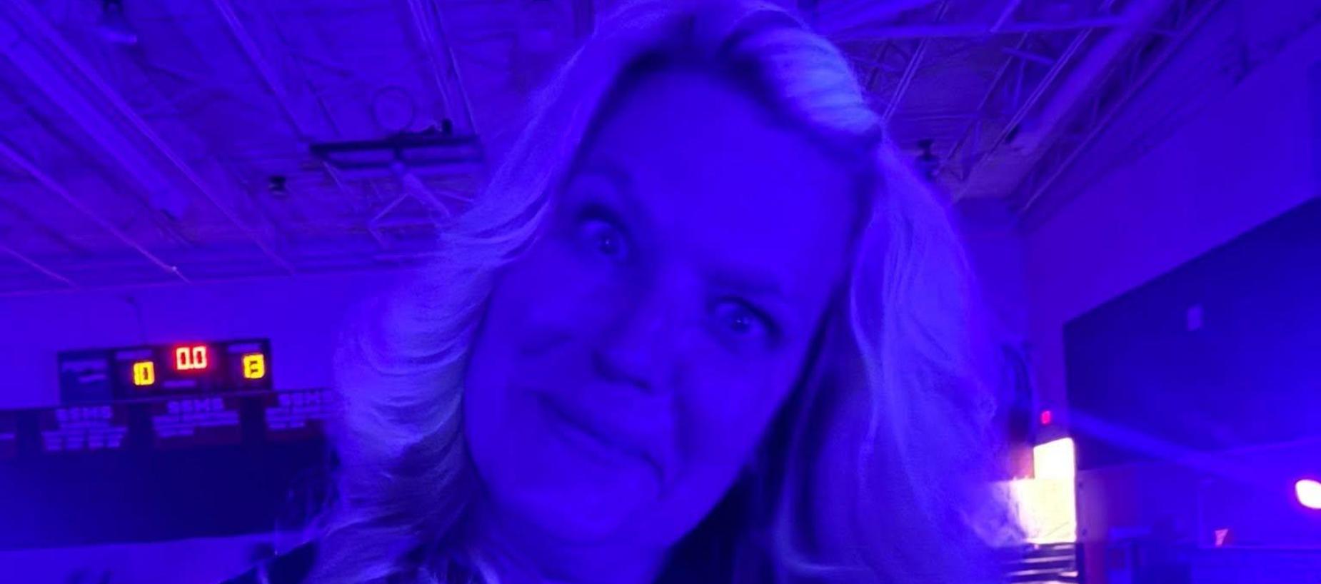 A person with long, wavy hair smiles under blue light in a gym setting.