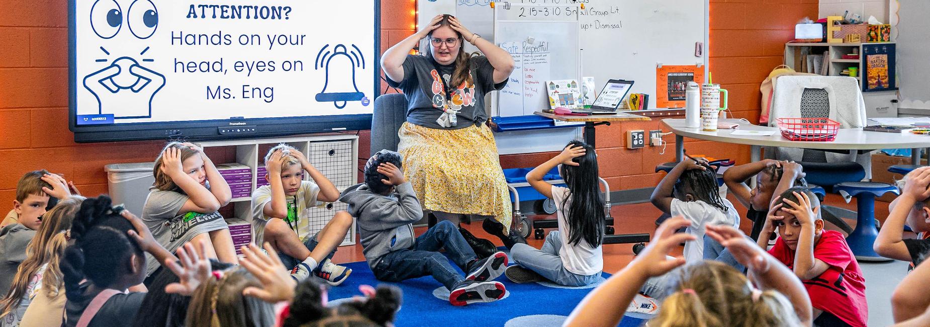 Students sitting on the floor attentively listening to their teacher in a colorful classroom.