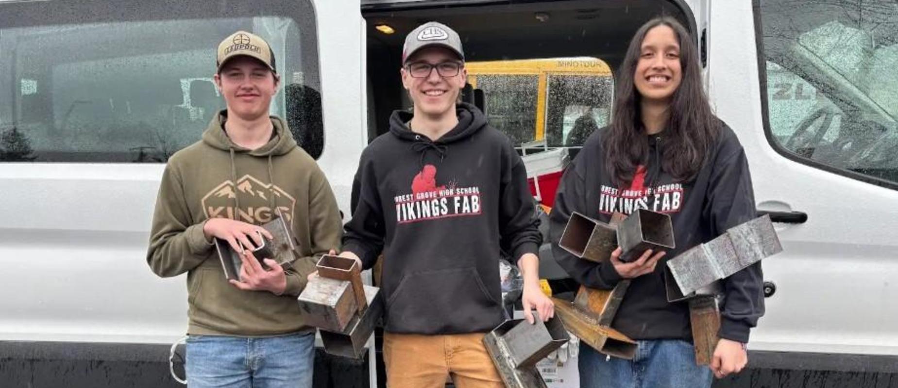 Three students hold metal pieces while standing in front of a van.