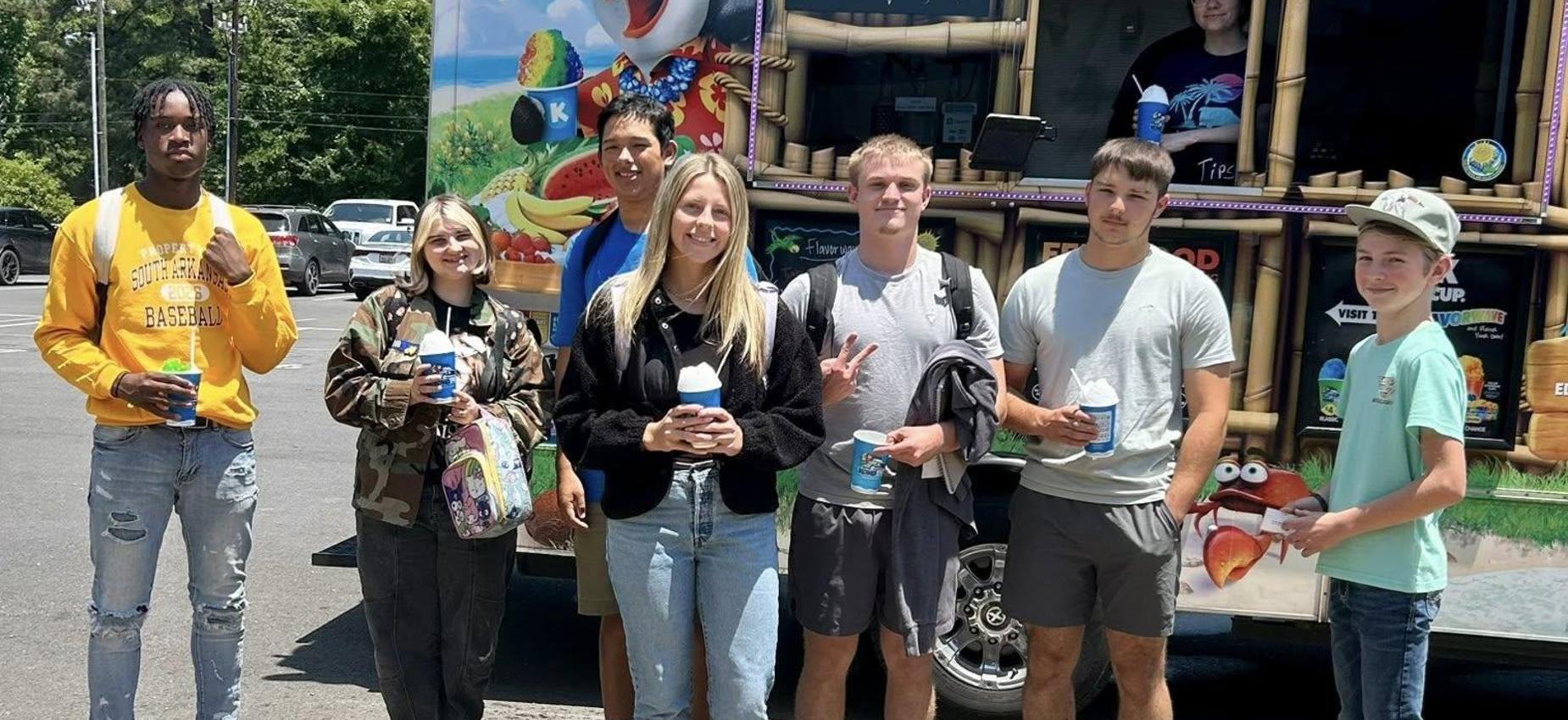 Group of seven young people posing with drinks near a colorful food truck.