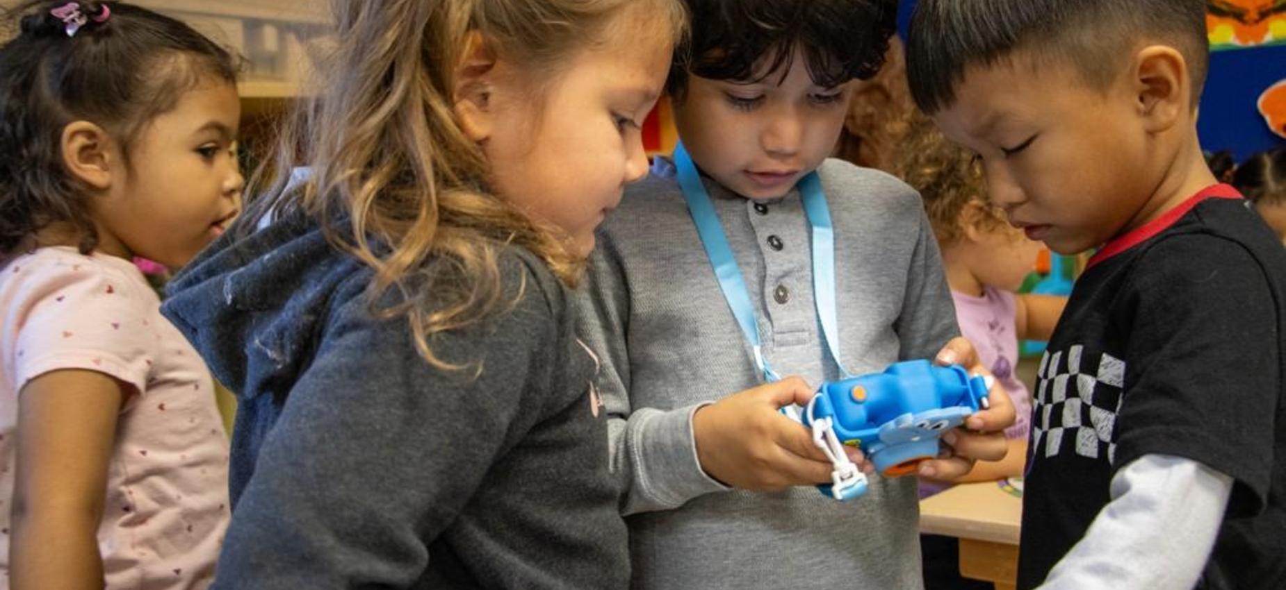 Three children focused on a toy camera while two others watch nearby.