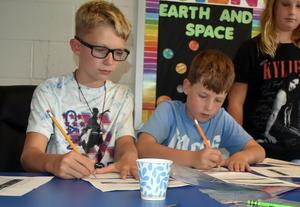 Two boys writing on papers at a table, with supplies and a cup visible.